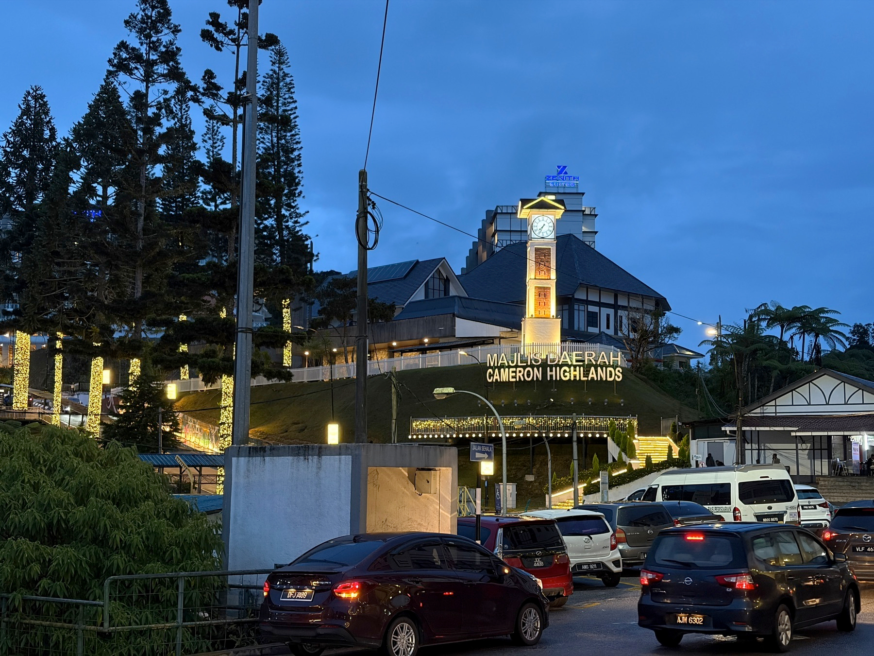 Auto-generated description: A clock tower and building in Cameron Highlands are illuminated at dusk with several cars parked nearby.