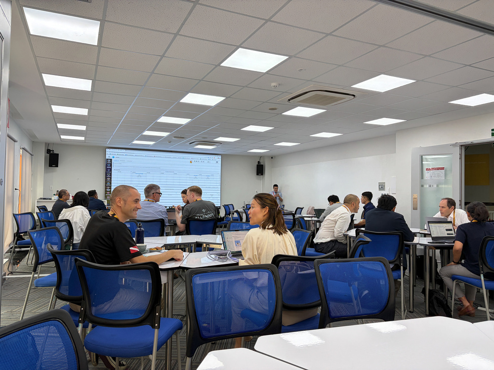 A group of people are seated on blue chairs in a modern classroom or training room, engaging in discussions with laptops and documents.