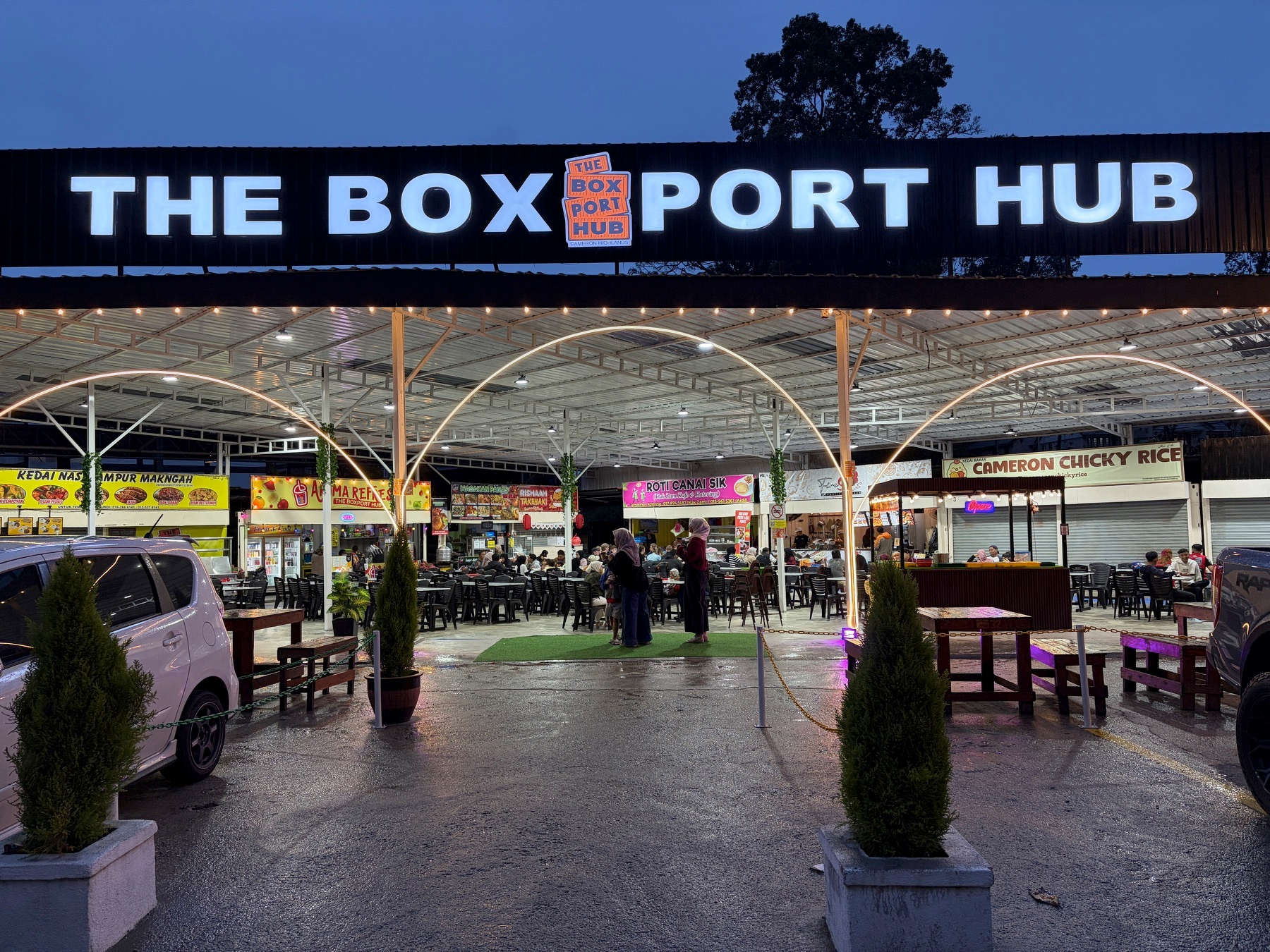 Auto-generated description: A lively food court area named The Box Port Hub is bustling with activity, featuring outdoor seating and various food stalls.