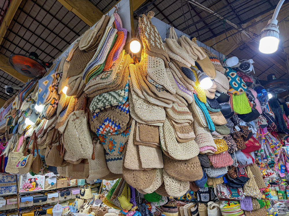 A market stall is displaying a wide variety of woven and crocheted bags and purses hanging closely together under bright lights.