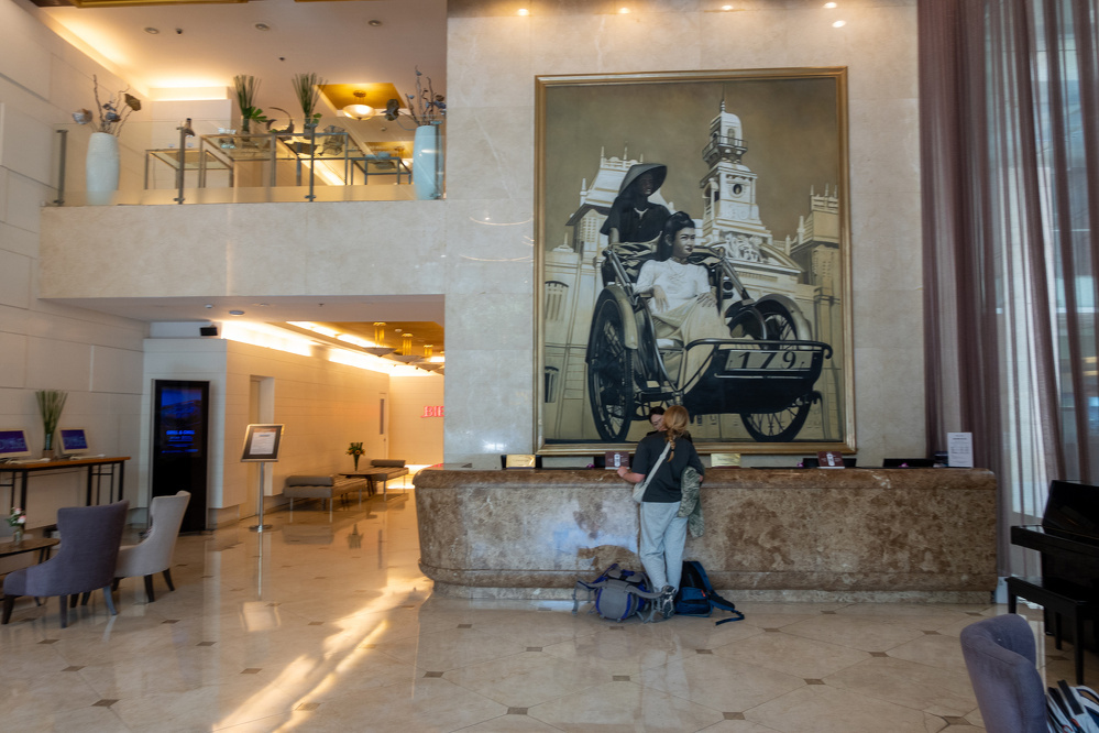 A person stands in a spacious, well-lit lobby with marble floors, large wall art depicting a vintage scene, and modern furnishings including chairs, tables, and decorative plants.