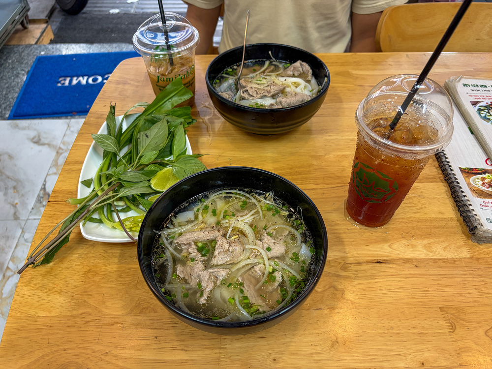 Two bowls of Vietnamese pho with herbs and lime, accompanied by iced beverages, are set on a wooden table at a restaurant.