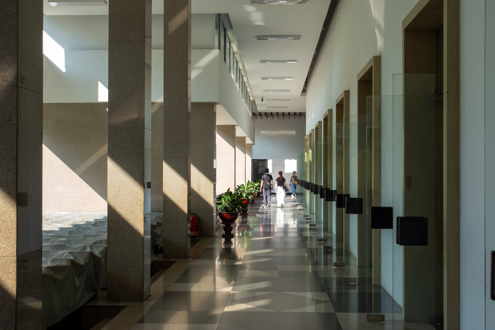 A bright corridor with glass-walled rooms on one side and large columns casting shadows on the floor, where a few people are walking in the distance.