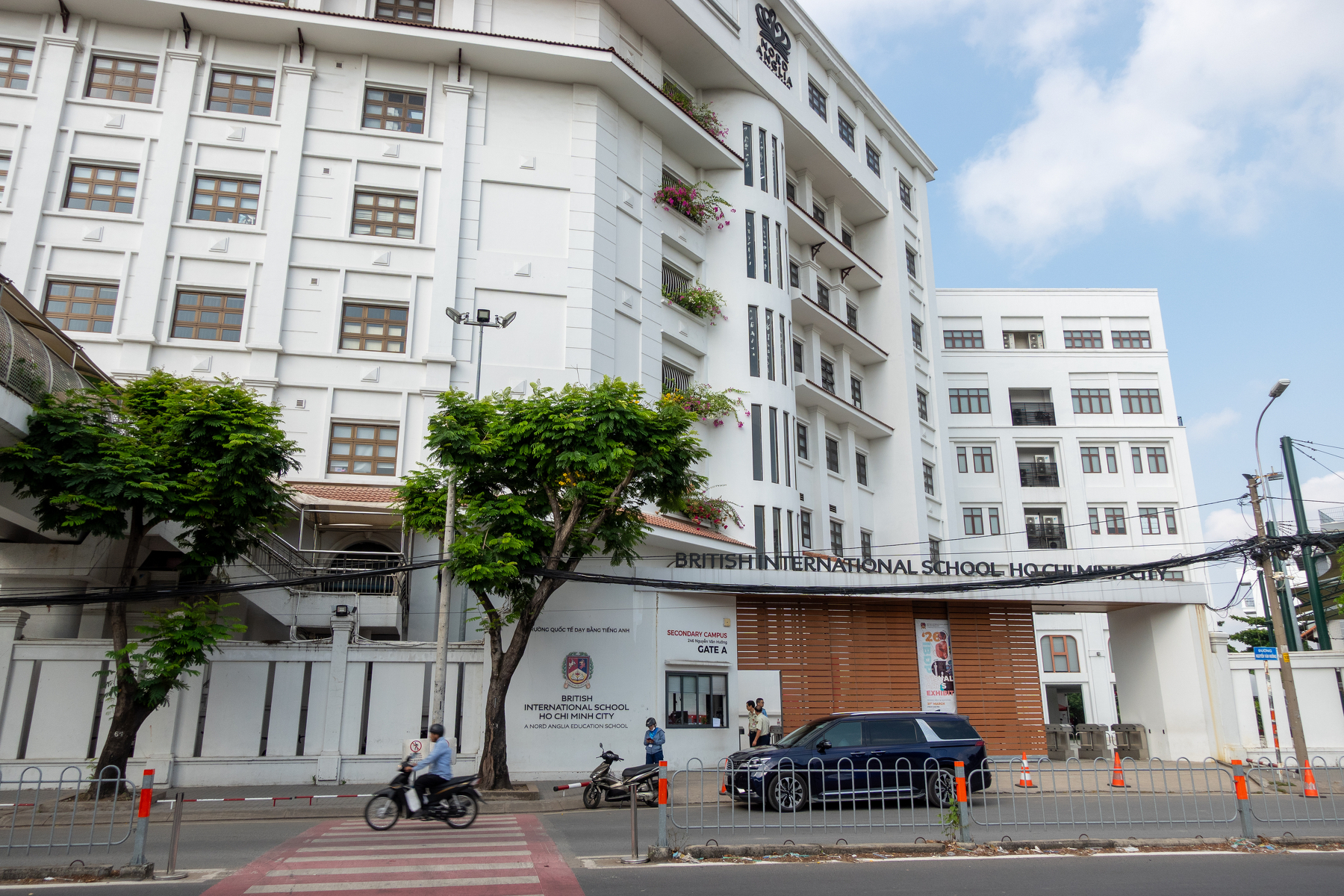A multi-story white building labeled British International School Ho Chi Minh is situated along a street with motorbikes and a car passing by.