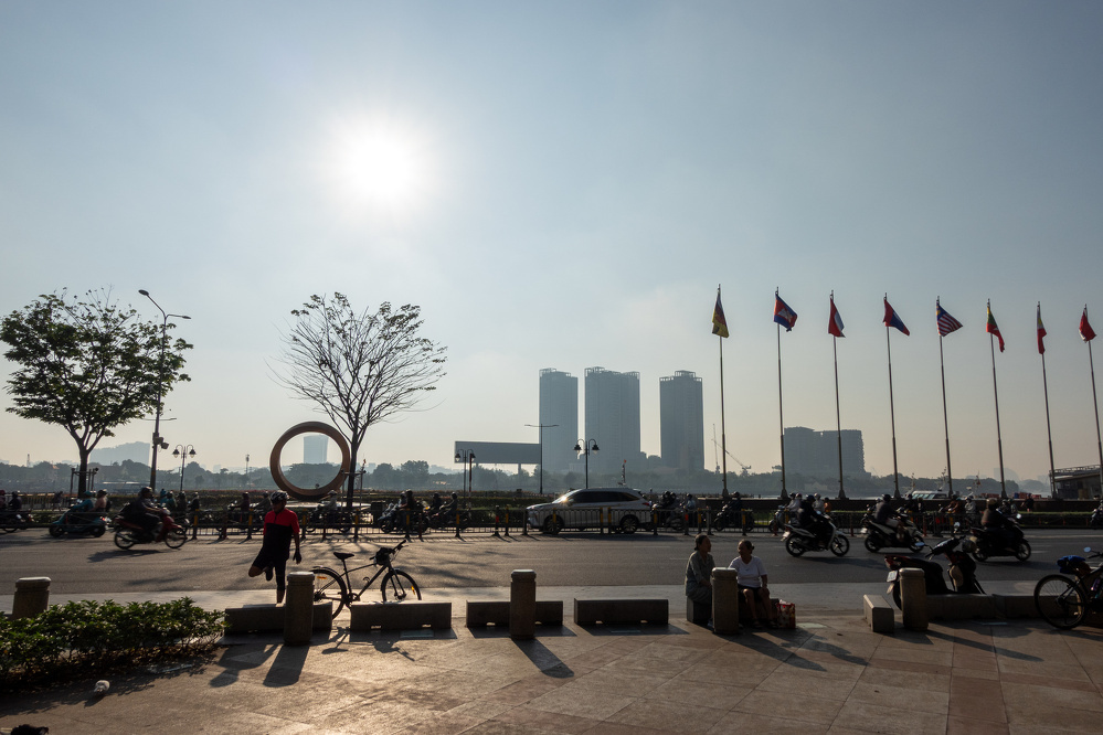 A busy street scene at sunset with people sitting near a row of flagpoles, motorcycles passing by, and tall buildings visible in the background.