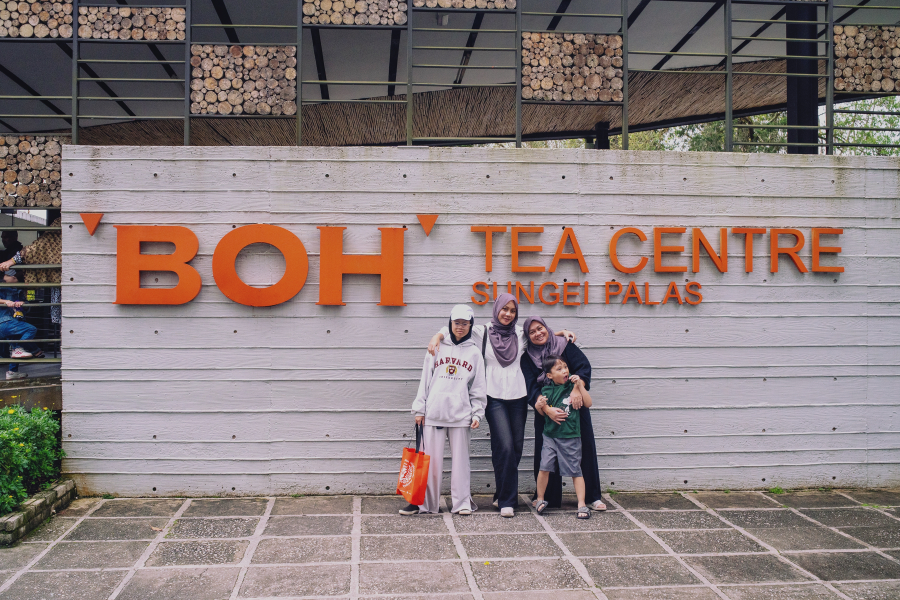 Auto-generated description: A group of people is posing in front of the BOH Tea Centre Sungai Palas sign with stacks of wooden logs above.