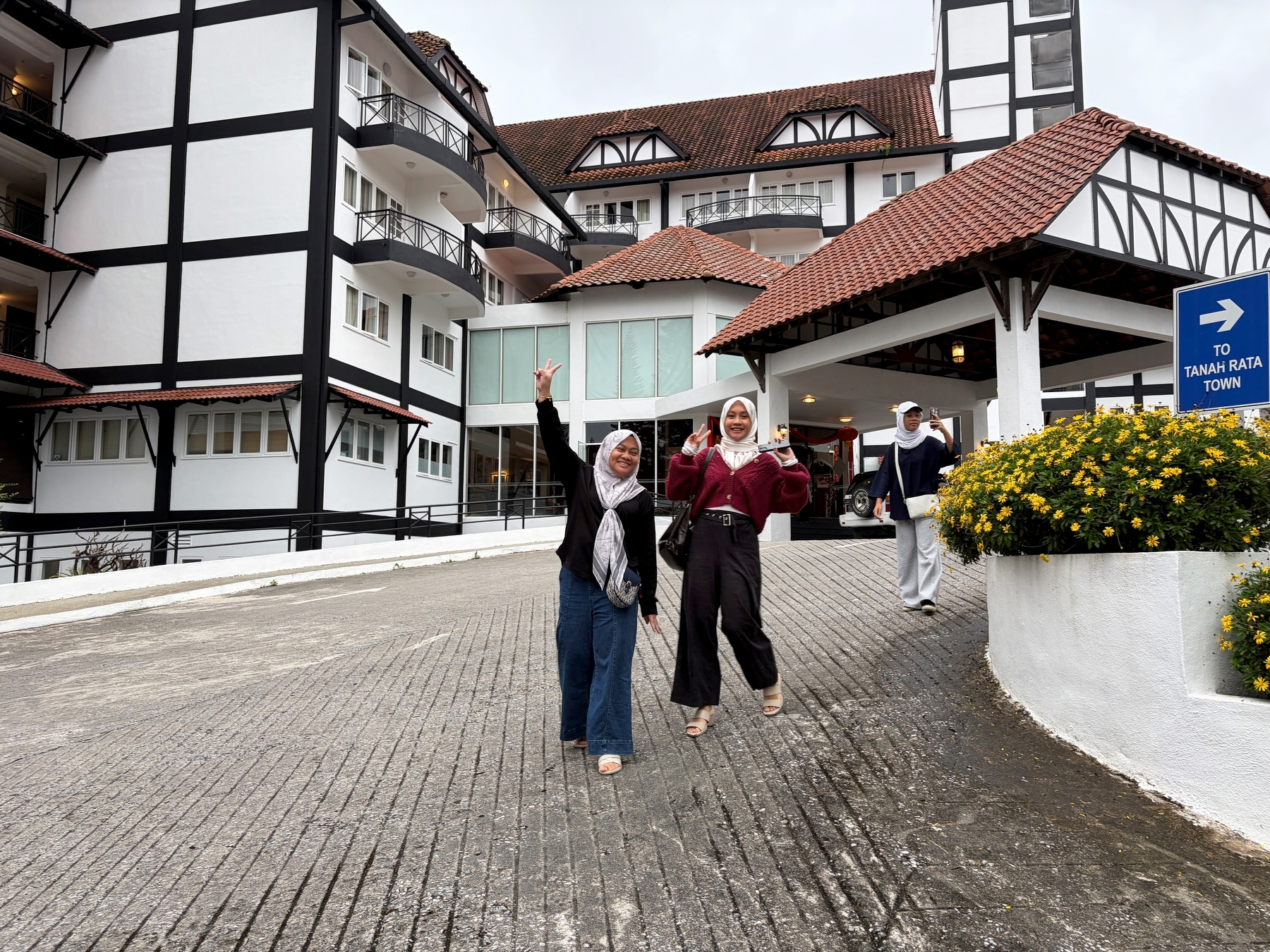 Auto-generated description: Two people pose cheerfully on a sloped path in front of a large, Tudor-style building with a person waving in the background.
