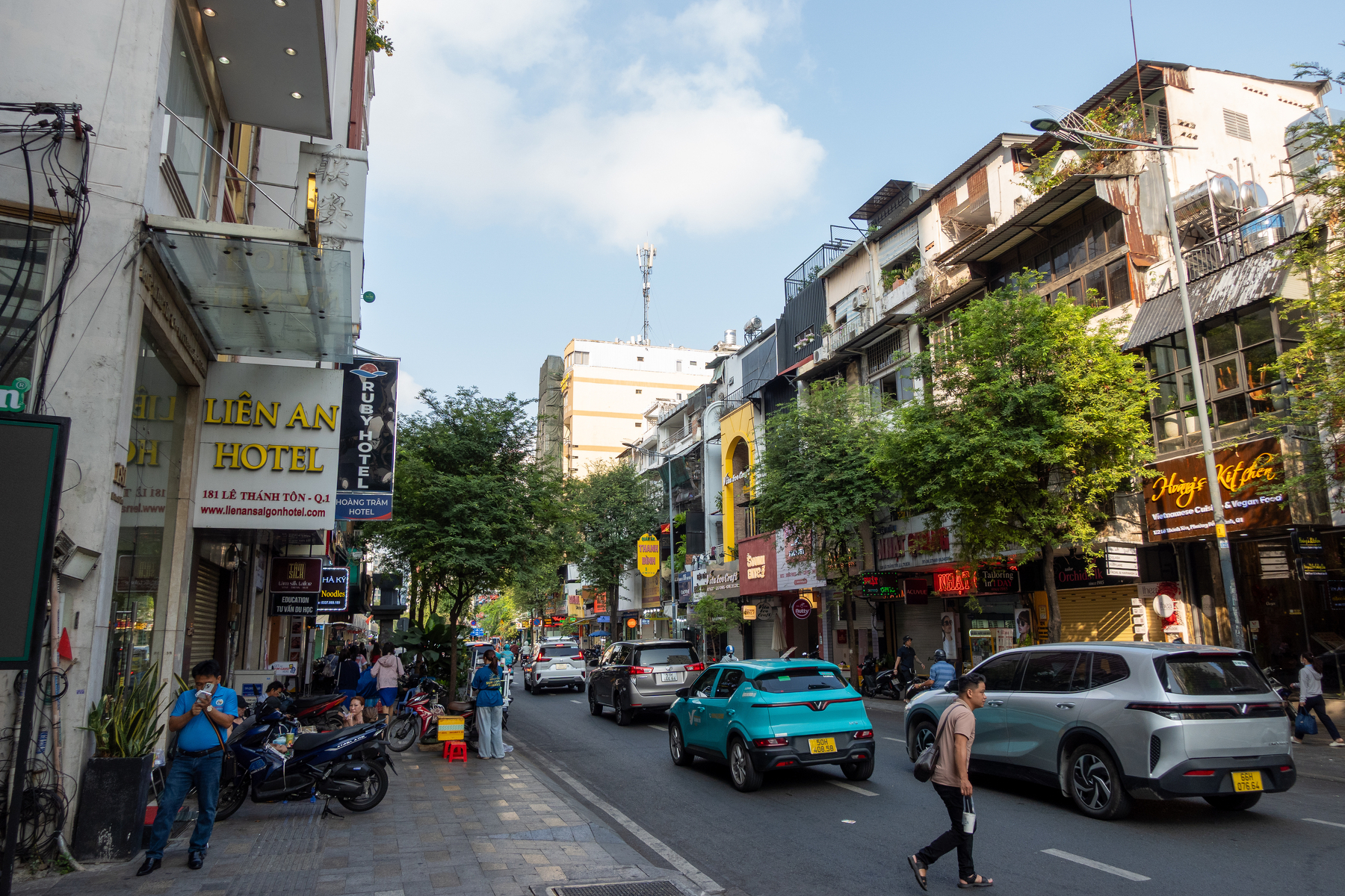 A busy urban street scene features cars, motorcycles, pedestrians, and various shops and hotels under a partly cloudy sky.
