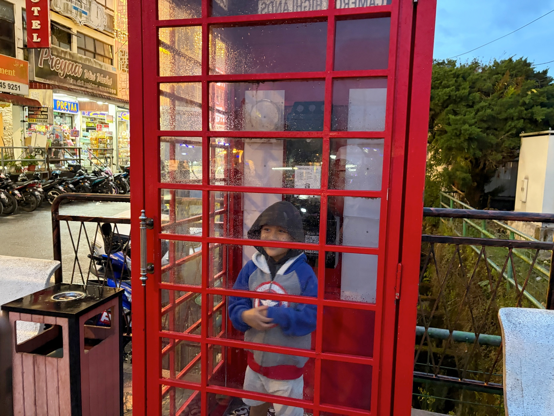Auto-generated description: A child is standing inside a red British-style telephone booth on a bustling street with various signs and motorcycles parked nearby.