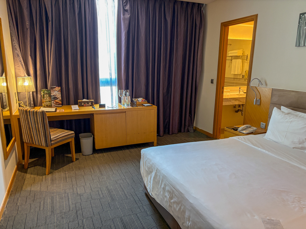 A clean and neatly arranged hotel room features a large bed, a wooden desk with a chair, and an open door leading to a bathroom.