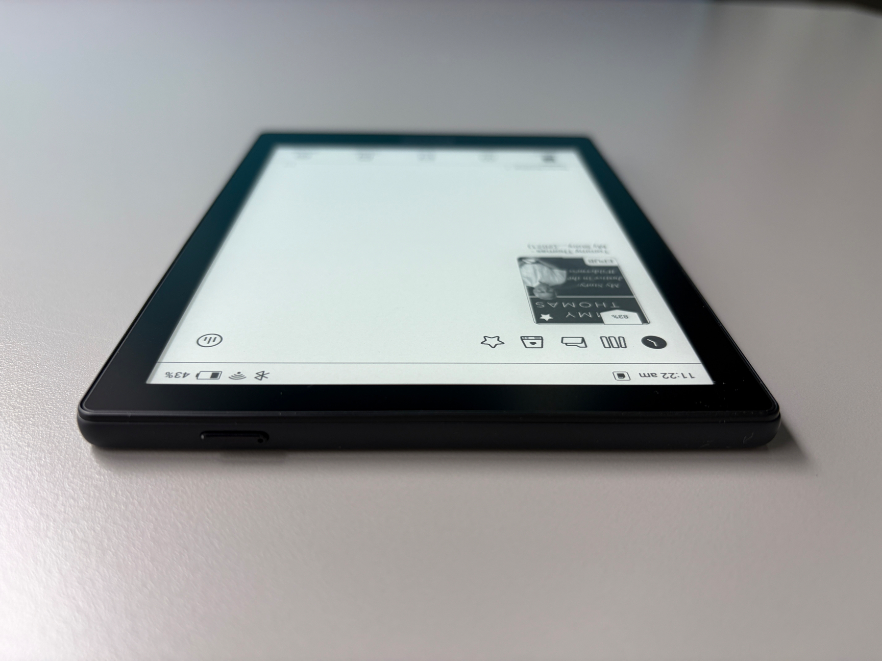Auto-generated description: A black e-reader device lies flat on a white surface displaying a page with mostly white space and a small image in the upper right corner.