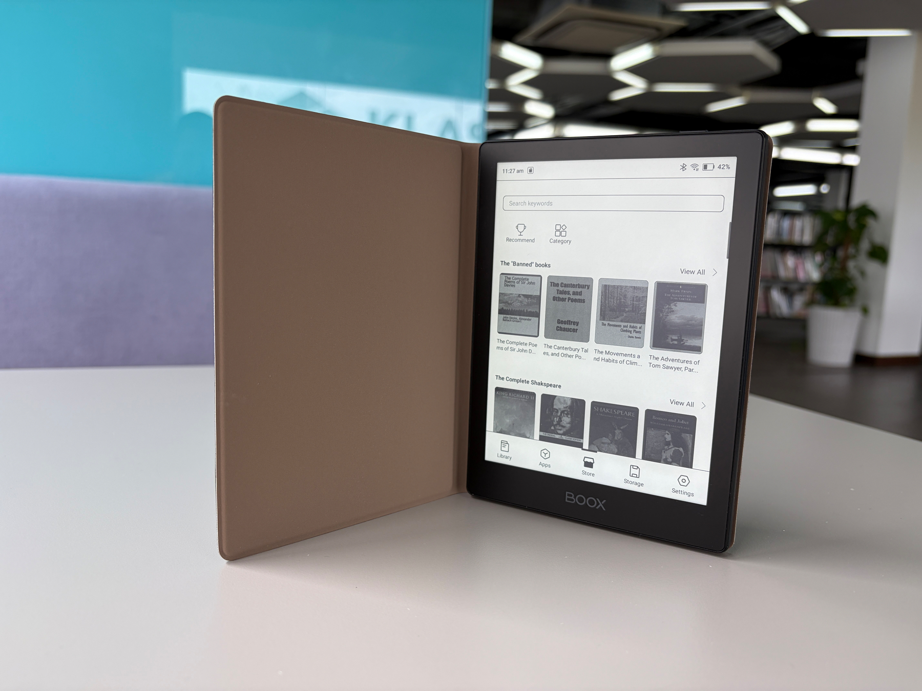 Auto-generated description: An e-reader displaying a digital bookshelf is propped open on a white table in a modern indoor setting.