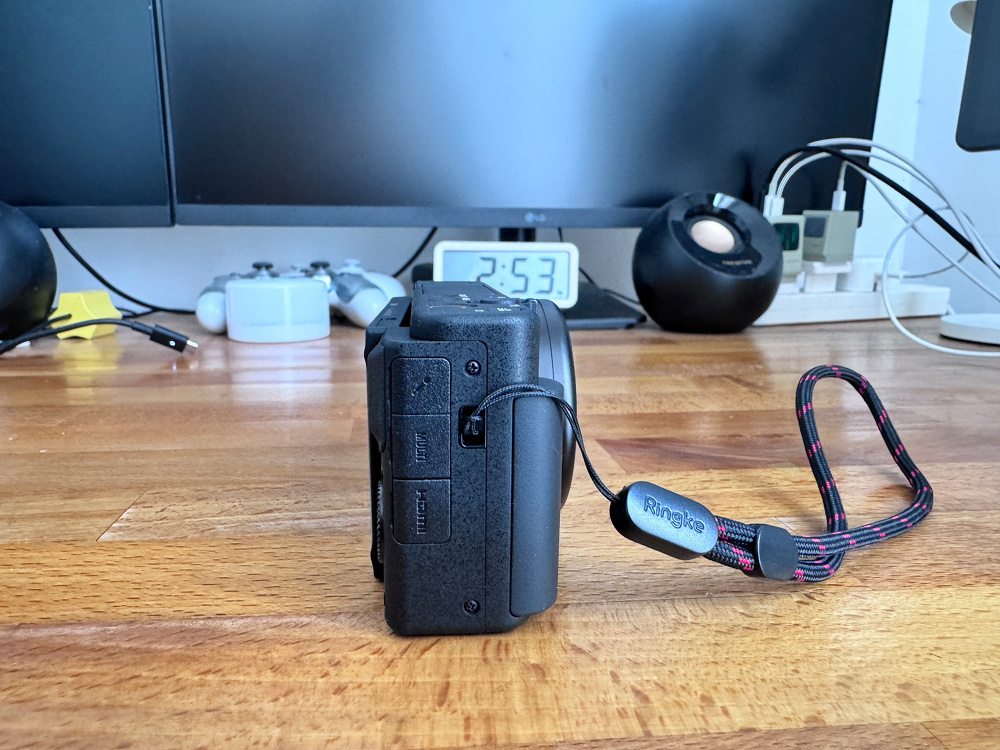 A camera attached to a shoulder strap is placed on a wooden desk in front of dual computer monitors and gaming controllers.