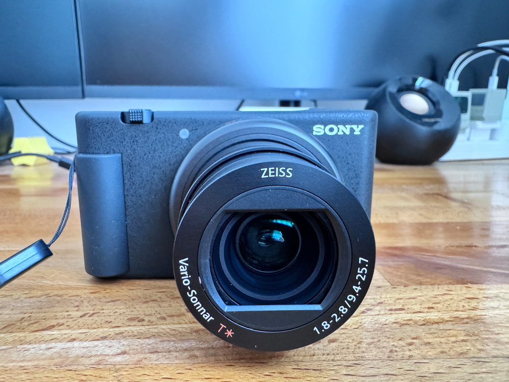 A Sony camera with a Zeiss lens rests on a wooden surface, with electronic equipment in the background.