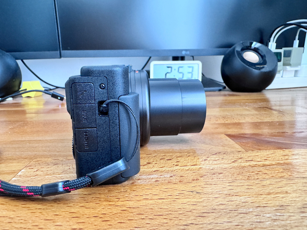 A camera with an extended lens is resting on a wooden desk in front of a digital clock displaying 2:57.