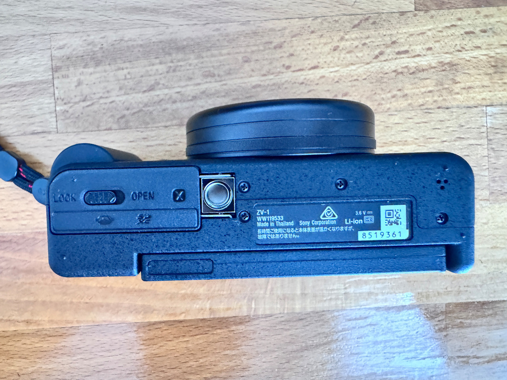 A digital camera is resting on a wooden surface with its bottom side visible, displaying various labels and the battery compartment.