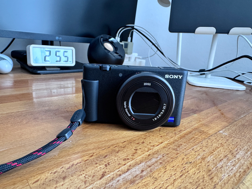 A Sony camera with a Zeiss lens is placed on a wooden desk, surrounded by a computer monitor, a clock displaying 2:55, and various electronic accessories.