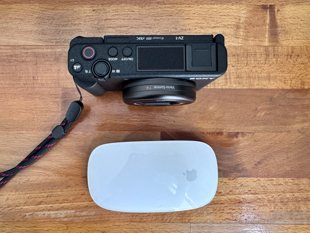 A camera with a lens and a wrist strap is placed above an Apple Magic Mouse on a wooden surface.