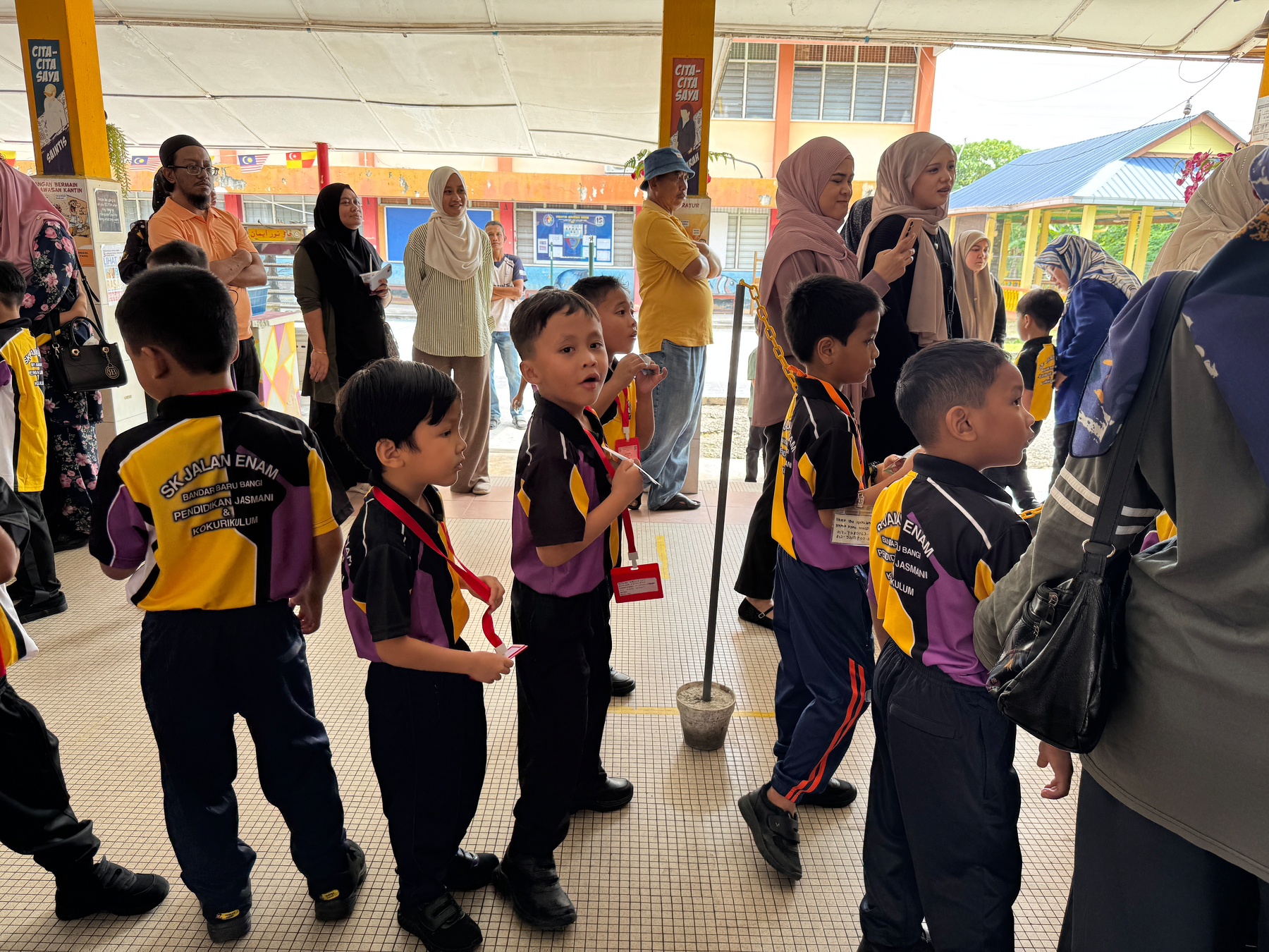 Auto-generated description: A group of young children in matching uniforms stand in line with adults, possibly teachers or parents, nearby.