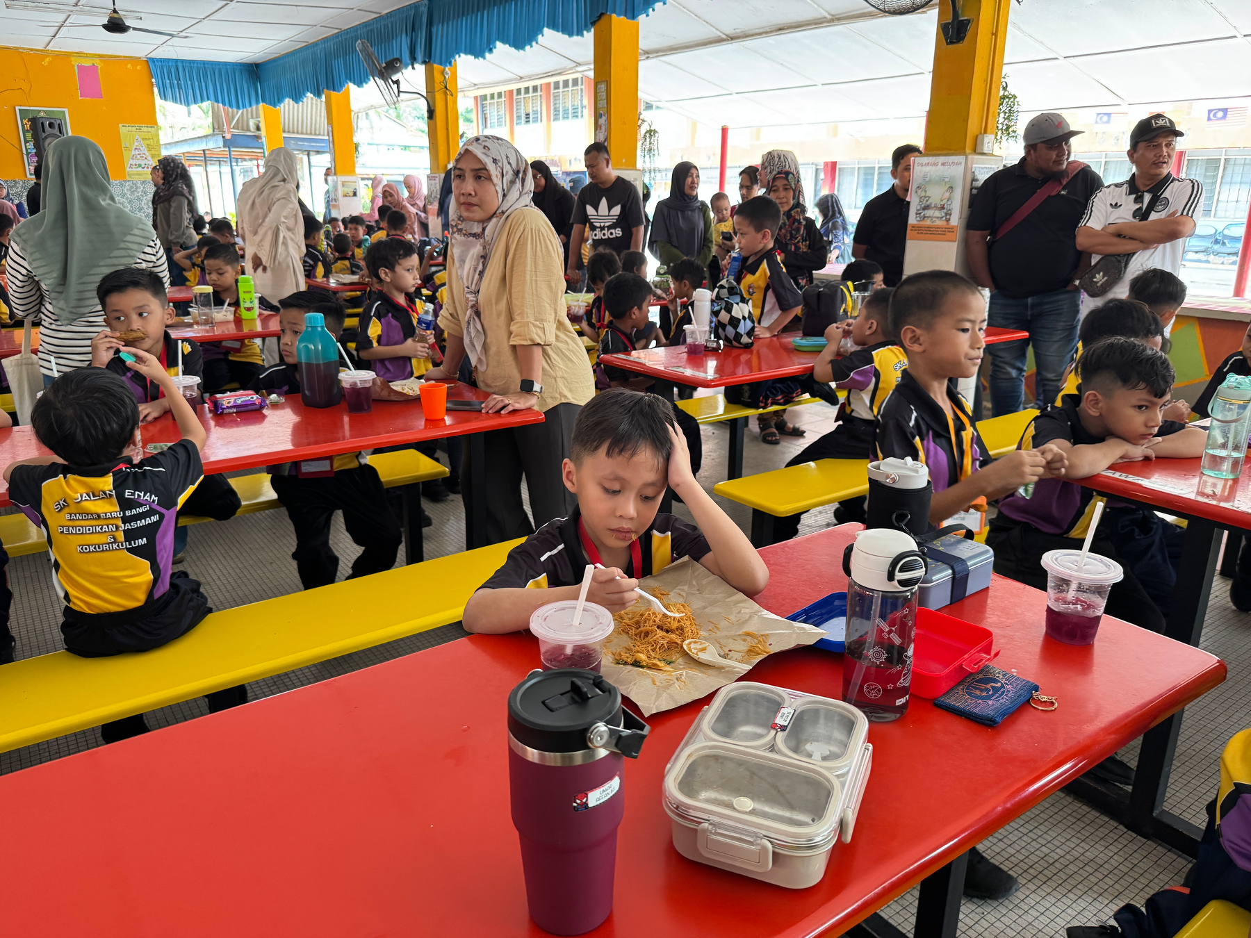 Auto-generated description: Children wearing uniforms are eating and sitting at red tables in a covered outdoor area, with a few adults standing nearby.