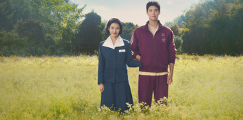 A young couple stands in a field of wildflowers, with the woman in a blue dress and the man in a maroon tracksuit, against a backdrop of trees and a blue sky.