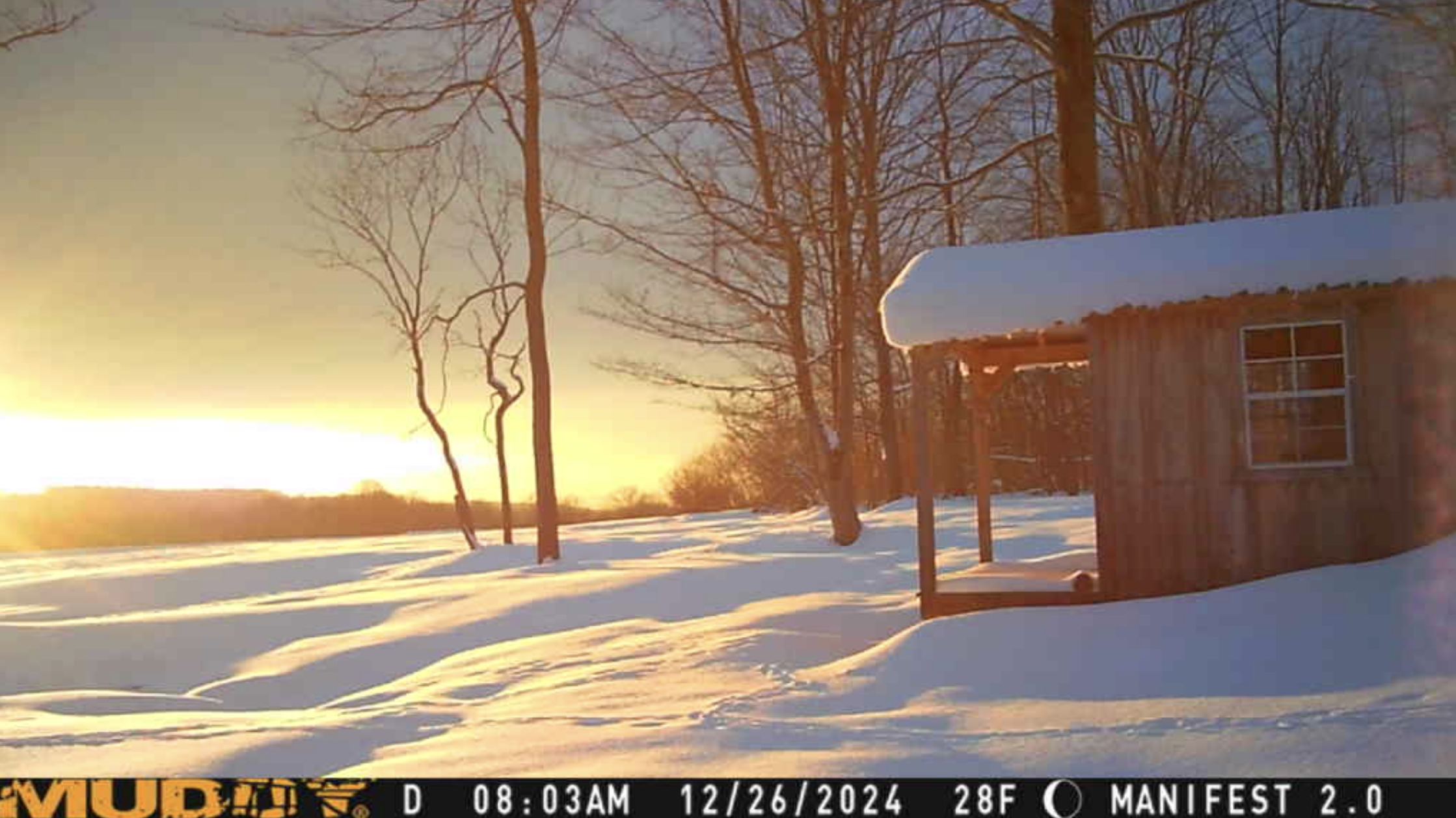 A snow-covered landscape features a small wooden cabin with a sunrise or sunset in the background.