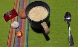 A mug of frothy beverage is surrounded by assorted treats and a spoon, all on a colorful striped and green placemat.