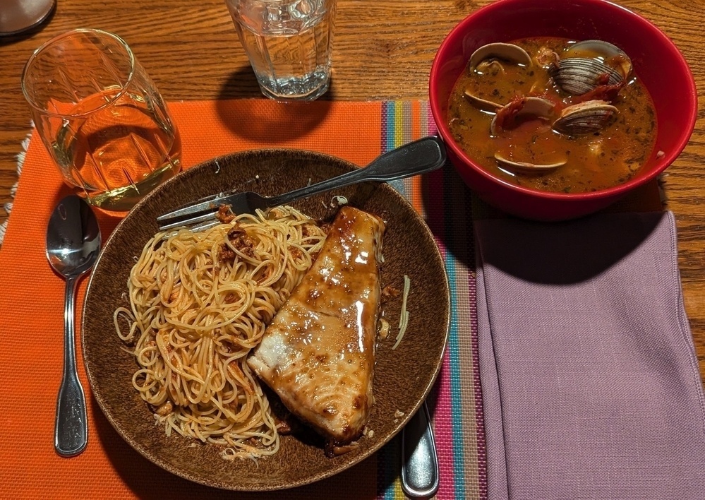 A plate of pasta with a grilled fish fillet and a bowl of clam soup are served on a colorful placemat with a glass of wine and water.