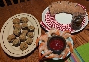 A table is set with a platter of stuffed mushrooms, a plate of shrimp with cocktail sauce, and sliced bread with a jar of spread.