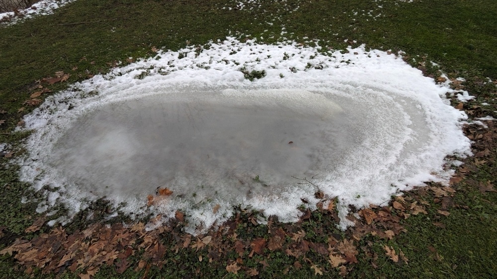 A shallow, icy puddle surrounded by snow and dead leaves on grass.