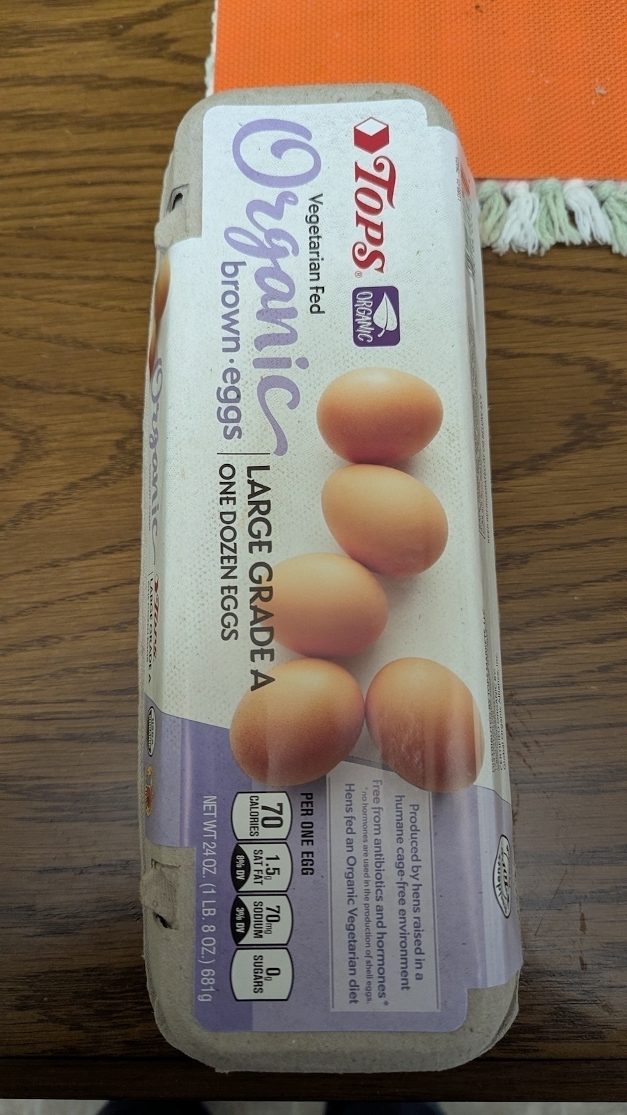 A carton of Tops Organic Vegetarian Fed Large Grade A brown eggs is placed on a wooden table.