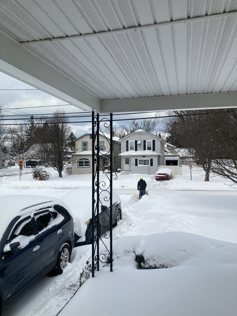 A person is walking down a snowy residential street lined with snow-covered cars and houses.