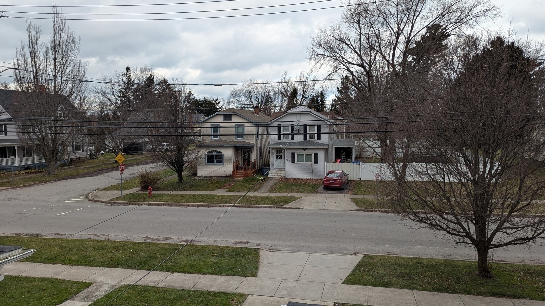 Auto-generated description: A suburban street is lined with houses and trees under a cloudy sky, with a parked red car near a white fence.