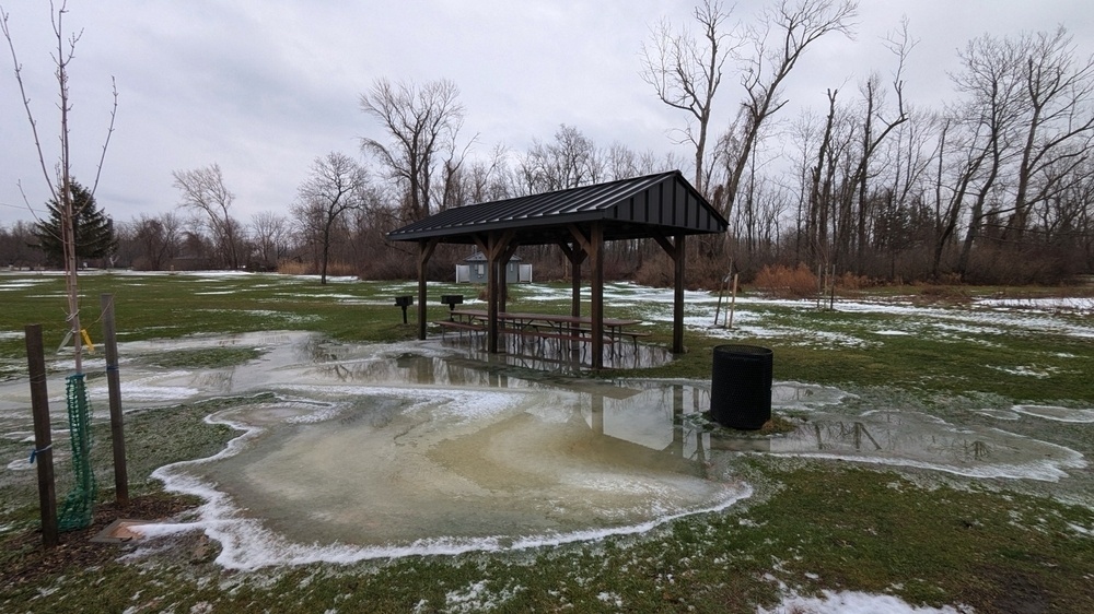 A picnic shelter stands surrounded by patches of ice and snow in a park with bare trees.