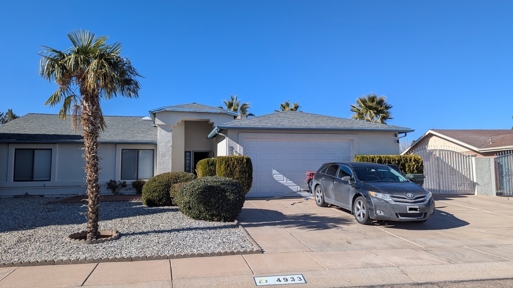 A single-story house with a two-car garage, a car parked in the driveway, and a few palm trees and shrubs in the yard under a clear blue sky.