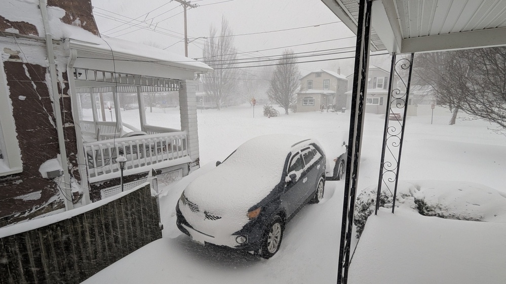 A car covered in snow is parked in a driveway during a heavy snowstorm, with snow accumulating on the surrounding area.