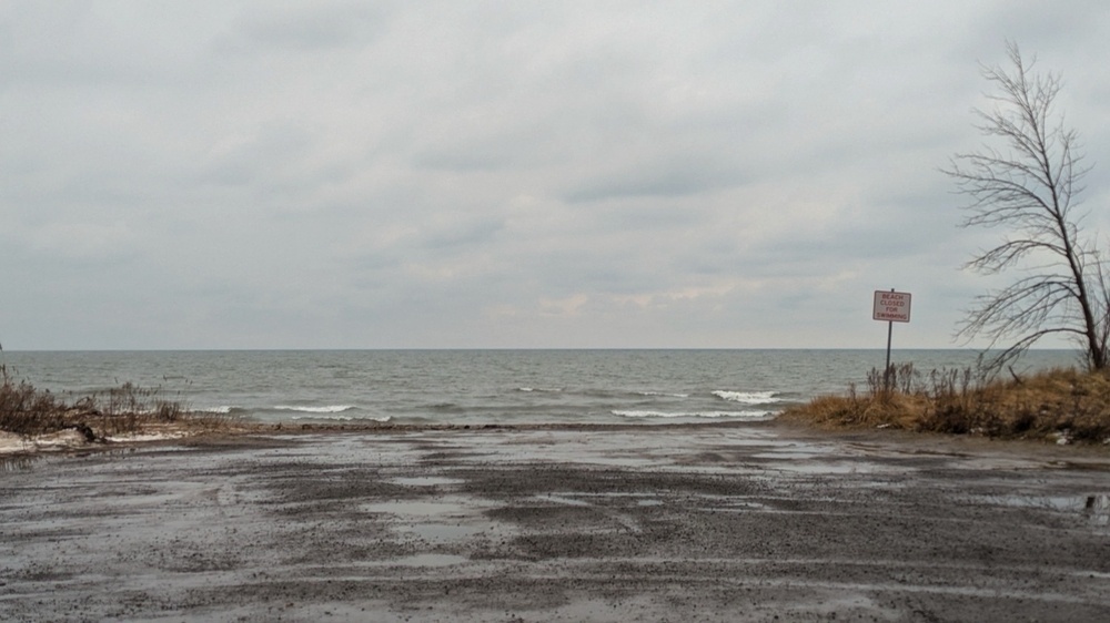 A cloudy day at a beach with a muddy path leading to the water, sparse vegetation, and a sign in the distance.