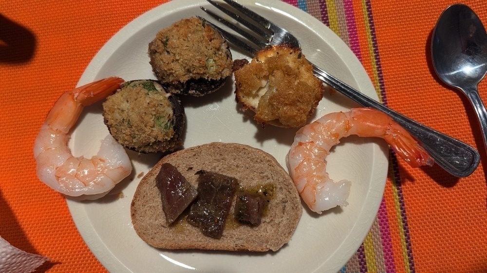 A plate of assorted appetizers, including shrimp, stuffed mushrooms, a bread slice with meat, and a small baked item, is set on a colorful tablecloth with utensils beside it.