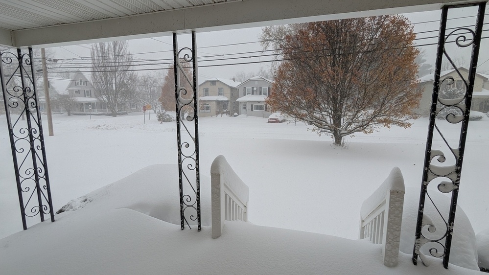 A snow-covered residential neighborhood is seen from a porch, with trees and houses blanketed in snow.