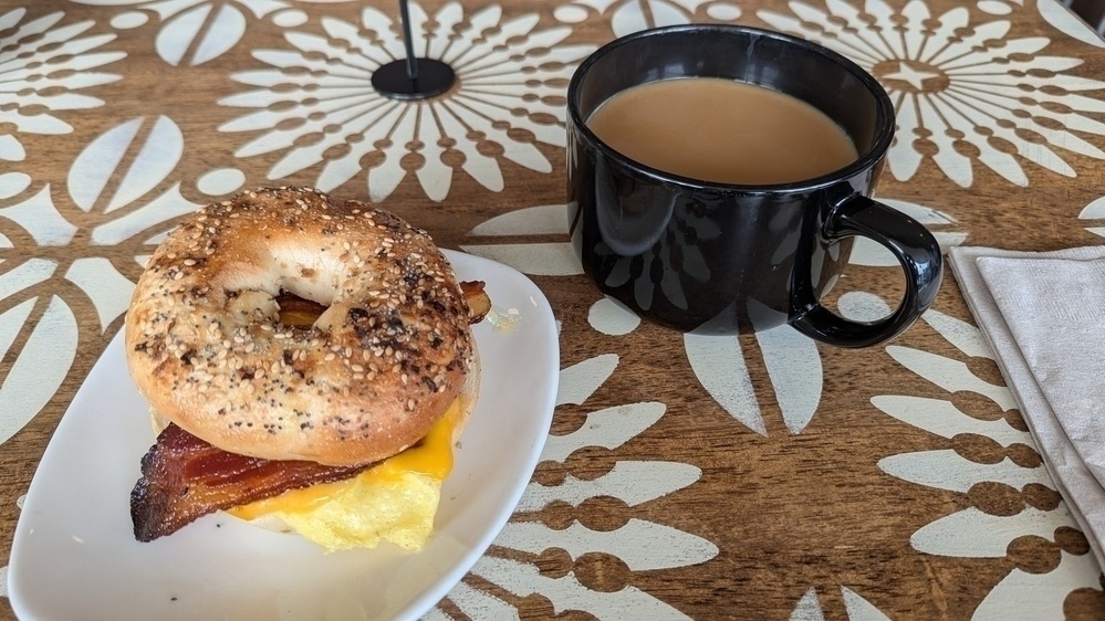 A bagel sandwich with bacon and cheese sits on a plate next to a black mug filled with coffee on a patterned table.