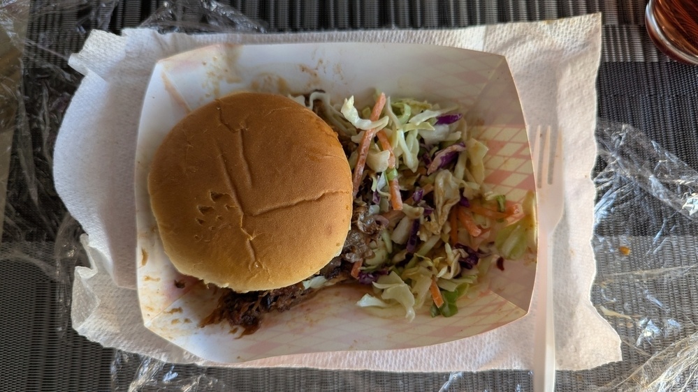 A pulled pork sandwich is served in a paper tray alongside a portion of coleslaw, with a plastic fork and a paper towel underneath.