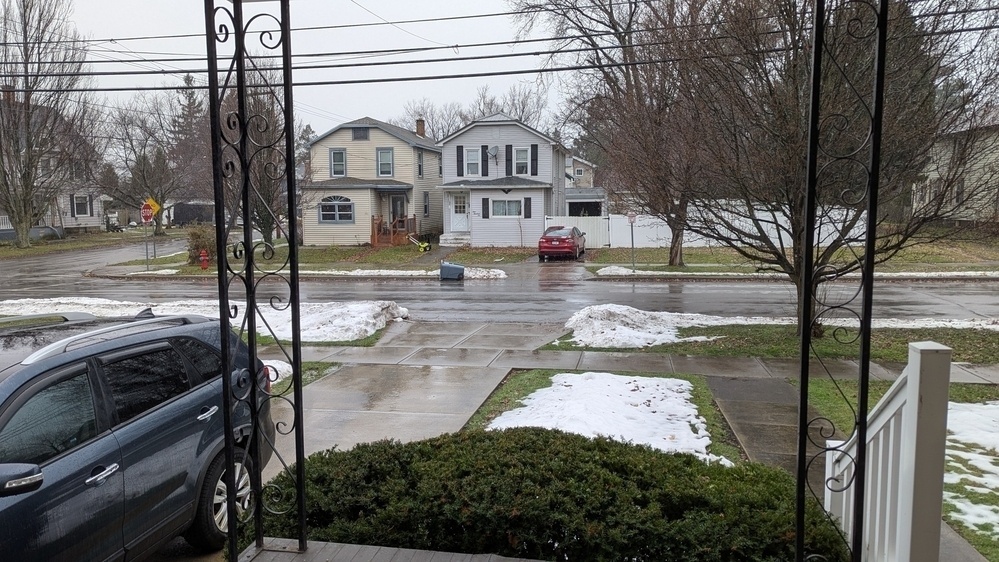 A suburban street is shown with parked cars, houses, and patches of melting snow on lawns and sidewalks.