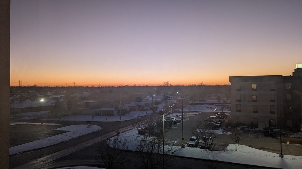 A quiet, snowy urban scene at dusk features a gradient sky transitioning from warm orange to deep blue, with buildings and streets bathed in the fading light.