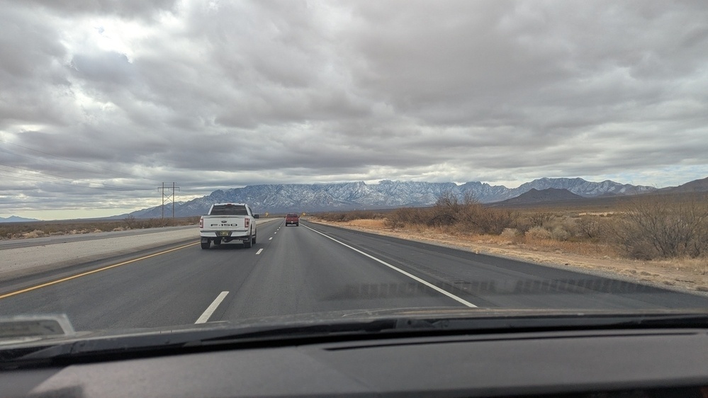 A stretch of highway with vehicles is flanked by desert terrain and distant mountains under a cloudy sky.