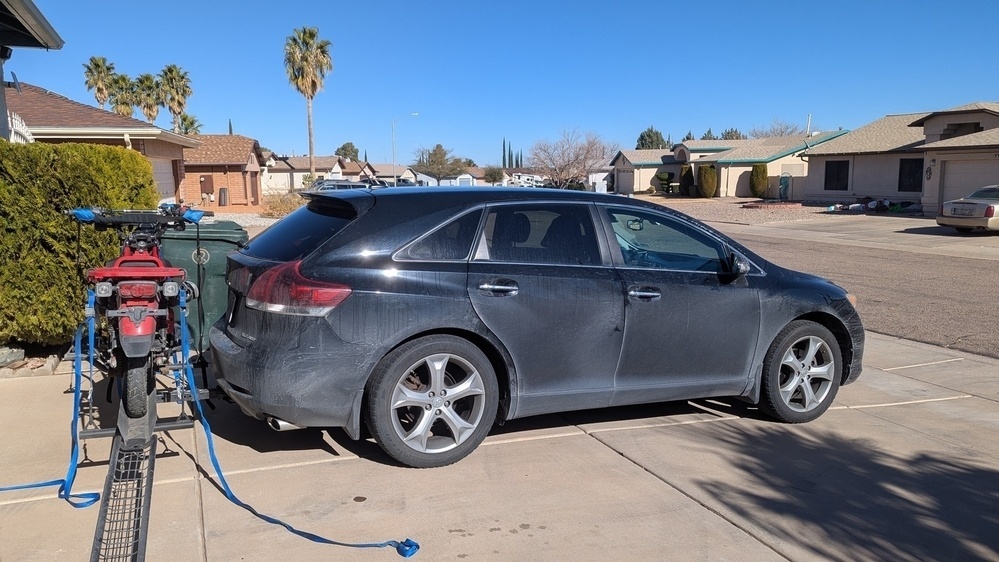 A black car with a bicycle mounted on its rear is parked in a driveway in a suburban neighborhood.