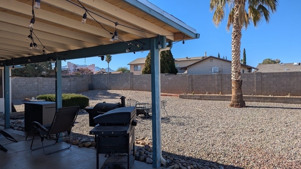 A backyard features a covered patio with furniture, a grill, and string lights, overlooking a gravel yard with a palm tree and a distant view of houses.