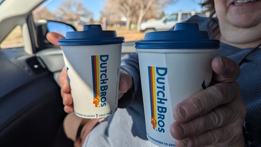 Two hands holding Dutch Bros coffee cups inside a car.