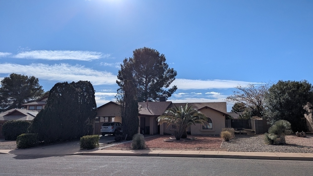 A suburban house is surrounded by trees and desert landscaping under a clear blue sky.