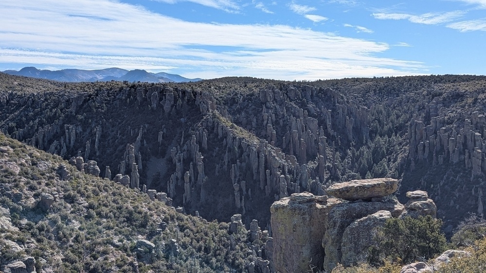 A rocky landscape features tall, jagged cliffs and scattered vegetation under a partly cloudy sky.