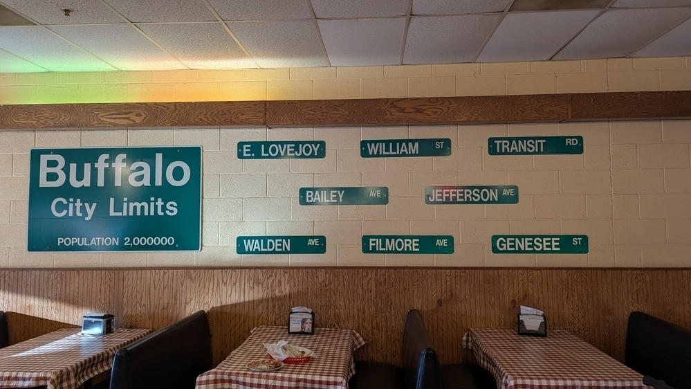 Several street signs are mounted on a wall inside a diner with checkered tablecloths on the tables.